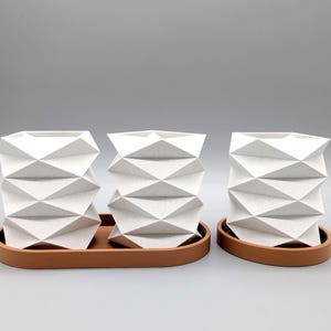 Könnte beinhalten: Drei weiße geometrische Pflanzgefäße mit strukturierter Oberfläche stehen auf einem terrakottafarbenen Tablett. Die Pflanzgefäße haben ein einzigartiges, eckiges Design. Das Tablett ist oval geformt. Der Hintergrund ist einfarbig hellgrau.