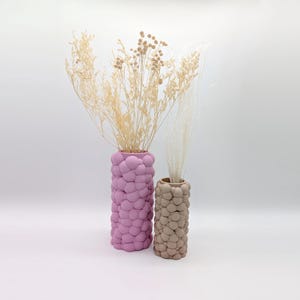 May include: Two decorative vases with a textured, bubble-like design. One vase is a dusty rose colour, and the other is a light beige. Both vases hold dried floral arrangements. The vases are cylindrical and stand upright.