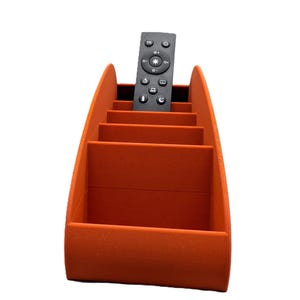 May include: An orange remote control tidy with several compartments. A black remote control sits inside the organiser. The tidy is made of a textured material and has a curved design.