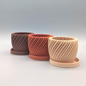 Könnte beinhalten: Drei dekorative Blumentöpfe mit Spiralstruktur in Braun-, Orange- und Beigetönen, jeweils auf einer passenden Untertasse. Die Töpfe haben ein modernes Design und eignen sich für kleine Pflanzen oder Sukkulenten.