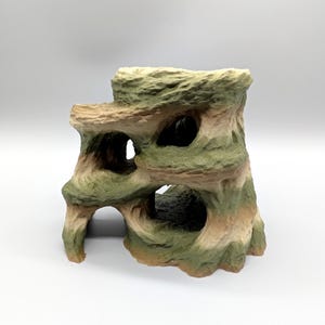 Peut inclure: Une structure en forme de roche à plusieurs niveaux présentant une surface texturée avec des bandes alternées de vert sourd et de beige. L'objet comprend plusieurs ouvertures creuses et des tunnels arqués créant un aspect caverneux.