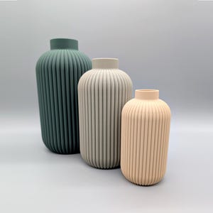 Puede incluir: Tres jarrones decorativos de diferentes tamaños y colores. El más grande es verde azulado, el del medio gris claro y el más pequeño melocotón. Cada jarrón tiene un diseño vertical acanalado.