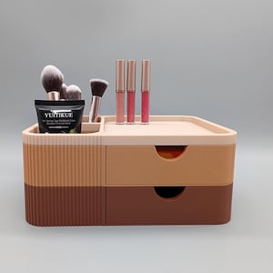Peut inclure: Un organisateur de maquillage bicolore avec deux tiroirs et un compartiment supérieur. L'organisateur est beige, beige clair et marron. Il contient des pinceaux de maquillage, des rouges à lèvres et un tube noir de masque facial YUITIKUE.