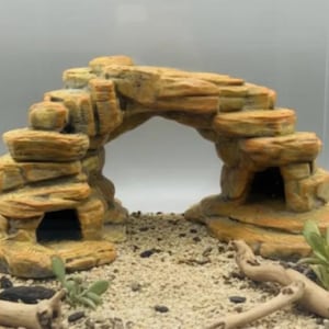 May include: A tan and brown rock-like aquarium ornament shaped as an arched bridge with two hollow cave entrances. The structure features layered, textured stone details and sits on a light-coloured sandy base with small pieces of driftwood.