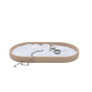Puede incluir: Una bandeja ovalada beige para joyas con un interior blanco texturizado, que contiene una pulsera plateada y varios anillos. El diseño de la bandeja se asemeja a un jardín zen, proporcionando una exhibición elegante para accesorios.
