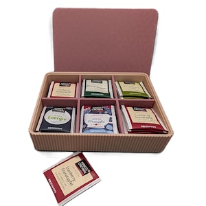 Könnte beinhalten: Eine hellrosa Schachtel mit verschiedenen Früchteteebeuteln. Die Schachtel ist in Fächer unterteilt, die jeweils eine andere Geschmacksrichtung enthalten. Die Beutel sind mit Texten wie "Cranberry-Granatapfel" und "Winter-Bratapfel" beschriftet.