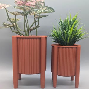 May include: Two terracotta-coloured planters with plants. The larger planter holds a plant with pink and green leaves. The smaller planter contains green grass-like foliage. Both planters have a ribbed design and are elevated on four legs.