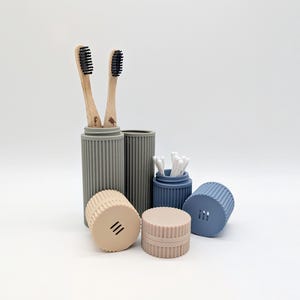 Op de afbeelding: Een verzameling badkameraccessoires in verschillende kleuren. Twee houten tandenborstels met zwarte borstelharen staan in een grijze houder. Andere containers zijn beige, blauw en grijs en bevatten wattenstaafjes en deksels. De items hebben een geribbeld ontwerp.