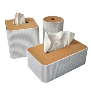 Pode incluir: Três caixas de lenços de papel com design moderno. As caixas são retangulares, quadradas e cilíndricas, com exterior cinza canelado e parte superior castanho-claro. Os lenços brancos são visíveis em cada abertura.