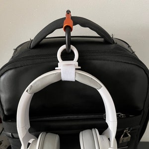 May include: White headphones with a black cord hanging from a black backpack. The headphones are attached to a white hook with a black carabiner clip.