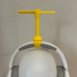 以下が含まれることがあります： グレーのヘッドバンドとイヤーカップが付いた白いヘッドセット。ヘッドバンドに十字形のトップが付いた黄色のプラスチックスタンドが取り付けられています。