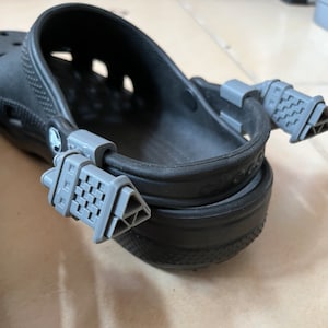 以下が含まれることがあります： グレーの調節可能なストラップが付いた黒いクロックスのペア。ストラップはテクスチャ加工されており、靴の後ろに付けられています。