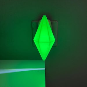May include: A green diamond-shaped night light plugged into an electrical outlet. The light is casting a green glow on the wall behind it.