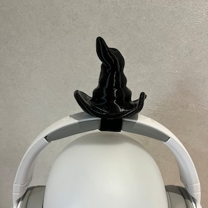 Pode incluir: Um chapéu de mago preto em miniatura para fones de ouvido. O chapéu está colocado em cima de um par de fones de ouvido brancos com fones de ouvido cinzentos. O chapéu é impresso em 3D e tem uma coroa pontiaguda e uma aba larga.