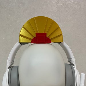 May include: A white headset with a gold and red headband. The headband is shaped like a sun with rays.