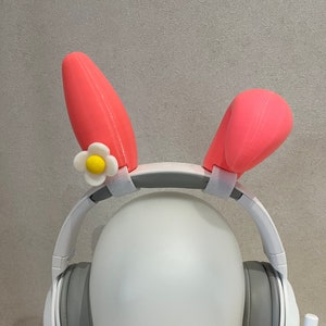 May include: White headphones with gray earcups and a pink bunny ear headband with a yellow flower accent.