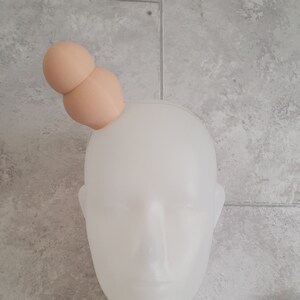 May include: A white mannequin head with a light brown headband with two round, light brown, fabric balls on top.