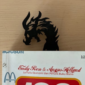 Puede incluir: Marcapáginas de dragón negro con un diseño detallado. El marcapáginas está encima de un libro con el texto "Emily Ross & Angus Holland, Jurnalis Ekonomi dan Penulis Buku Bisnis, 100"