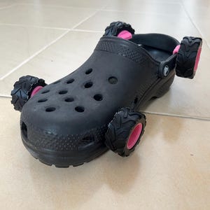 Puede incluir: Zueco negro modificado con cuatro ruedas negras y rosas de estilo todoterreno. El zapato tiene múltiples orificios de ventilación y una superficie texturizada. Las ruedas están unidas a los lados, creando un juguete único.