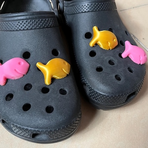 May include: A pair of black clogs decorated with fish-shaped charms in pink and gold. One clog has a pink and a gold fish, the other has a gold and a pink fish. These charms add a fun element to the casual shoes.