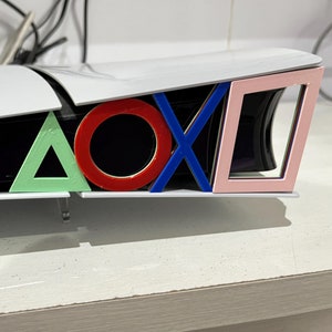 May include: A white PlayStation 5 console with a custom design featuring the PlayStation logo symbols in different colors: a green triangle, a red circle, a blue X, and a pink square.