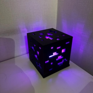 Può includere: Lampada nera a forma di cubo con intagli pixelati, che emette una vivace luce viola. La lampada è posizionata su una superficie bianca. Il design suggerisce un tema di videogioco.