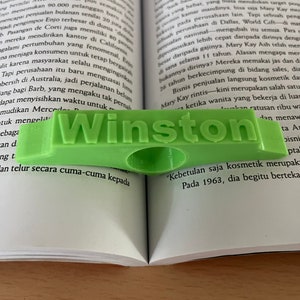 Op de afbeelding: Een groene plastic bladwijzer met het woord "Winston" erop gedrukt. De bladwijzer ligt op een open boek.