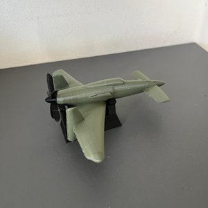 Può includere: Un modello di aereo piccolo di colore grigio stampato in 3D con un'elica nera. L'aereo è seduto su un supporto nero.