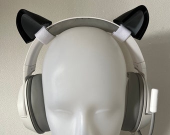 Puede incluir: Auriculares inalámbricos blancos con orejas de gato negras unidas a la diadema. Los auriculares están en una cabeza de maniquí blanca.