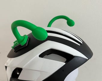 May include: A white and black bicycle helmet with green antennae attached. The helmet has the brand name "ROCKBROS" printed on the side. The antennae are curved and have rounded tips.