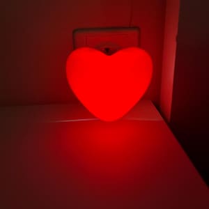 Könnte beinhalten: Ein rotes herzförmiges Nachtlicht, das in eine Steckdose gesteckt ist.