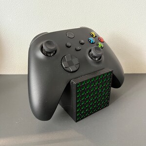 Può includere: Un controller Xbox nero è appoggiato su un supporto di ricarica nero con un motivo a griglia verde.