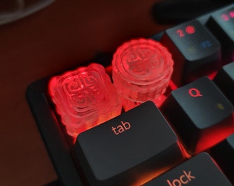 May include: Two clear red glowing keycaps with a design on top, sitting on a black keyboard with the keys "tab" and "caps lock" visible.