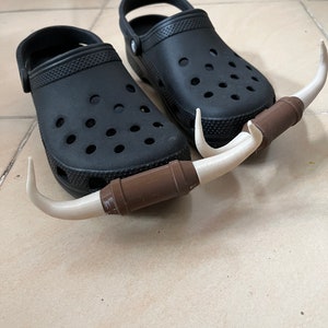 May include: Two black clogs with white horns attached to the front. The horns are made of a brown and white material.