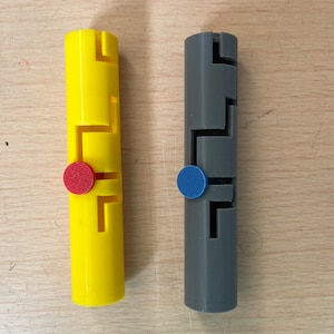 Könnte beinhalten: Zwei Puzzleteile, eines gelb und eines grau, mit einem roten und blauen Knopf auf jedem. Die Puzzleteile sind aus Kunststoff und haben ein komplexes Design.