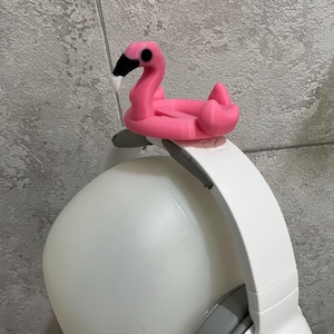 以下が含まれることがあります： 白いヘッドホンにピンクのフラミンゴの形をした携帯電話ホルダーが取り付けられています