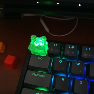 Puede incluir: Un tapón de tecla verde brillante con la letra 'G' en él, sentado en un teclado mecánico negro con retroiluminación arcoíris. El teclado tiene teclas etiquetadas 'tab', 'bloq mayús', 'shift', 'Q', 'W', 'A', 'S', 'Z', 'X', '1!', '2@', '3#', 'F1', 'F2', 'F3' y '80'.