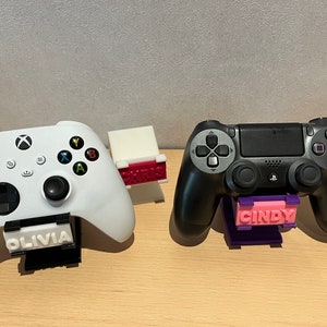 Può includere: Due controller per videogiochi, uno bianco e uno nero, sono esposti su supporti personalizzati. Il controller bianco ha un supporto con il nome "Olivia" stampato su di esso, e il controller nero ha un supporto con il nome "Cindy" stampato su di esso.