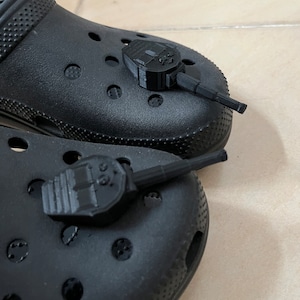 Op de afbeelding: Twee zwarte plastic tankcharms bevestigd aan een paar zwarte klompen. De tankcharms zijn gedetailleerd met een torentje en een loop.