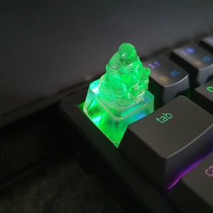 May include: A green, translucent, resin figurine of a seated Buddha on a keyboard keycap. The keycap is illuminated with green light. The keycap is labeled "tab" and is on a black mechanical keyboard.