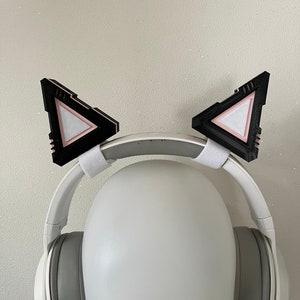 Puede incluir: Auriculares blancos con orejas de gato negras unidas a la diadema. Las orejas de gato tienen un diseño triangular rosa y blanco.