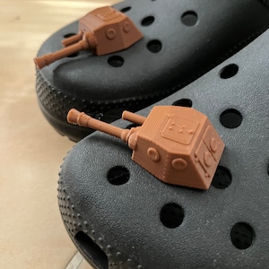May include: Two brown plastic toy tanks with black barrels are attached to a pair of black clogs.