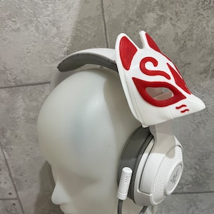 Könnte beinhalten: Weißes Gaming-Headset mit einem grauen Kopfbügel und einer weißen und roten Fuchsmaske, die oben befestigt ist. Das Headset hat ein Mikrofon, das am linken Ohrhörer befestigt ist.