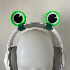 May include: White headphones with green frog-shaped ear covers. The ear covers have black eyes and white pupils.