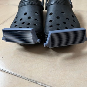 May include: A pair of black clogs with grey rubber treads attached to the bottom. The treads have a ridged design.