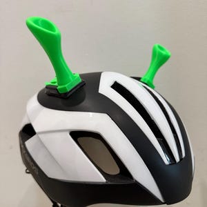 May include: A white and black bicycle helmet with green, horn-shaped attachments. The helmet features a streamlined design with black vents and a chin strap. The bright green attachments add a playful element to the helmet.