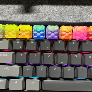 May include: A keyboard with rainbow gummy bear keycaps. The keycaps are translucent and show the rainbow colors underneath. The keyboard has a black frame and gray keys.