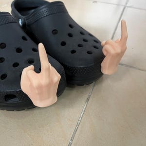 May include: A pair of black clogs with two small, flesh-colored, hand-shaped charms attached to the straps. The charms are making the middle finger gesture.