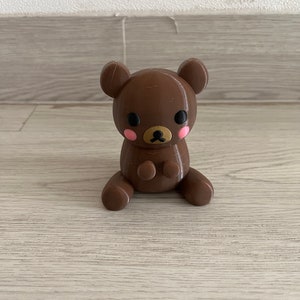 以下が含まれることがあります： 黒い目とピンクの頬をした茶色のぬいぐるみ。熊は前足を前に出して座っています。