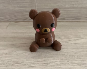 May include: A brown teddy bear with black eyes and pink cheeks. The bear is sitting with its paws in front of it.